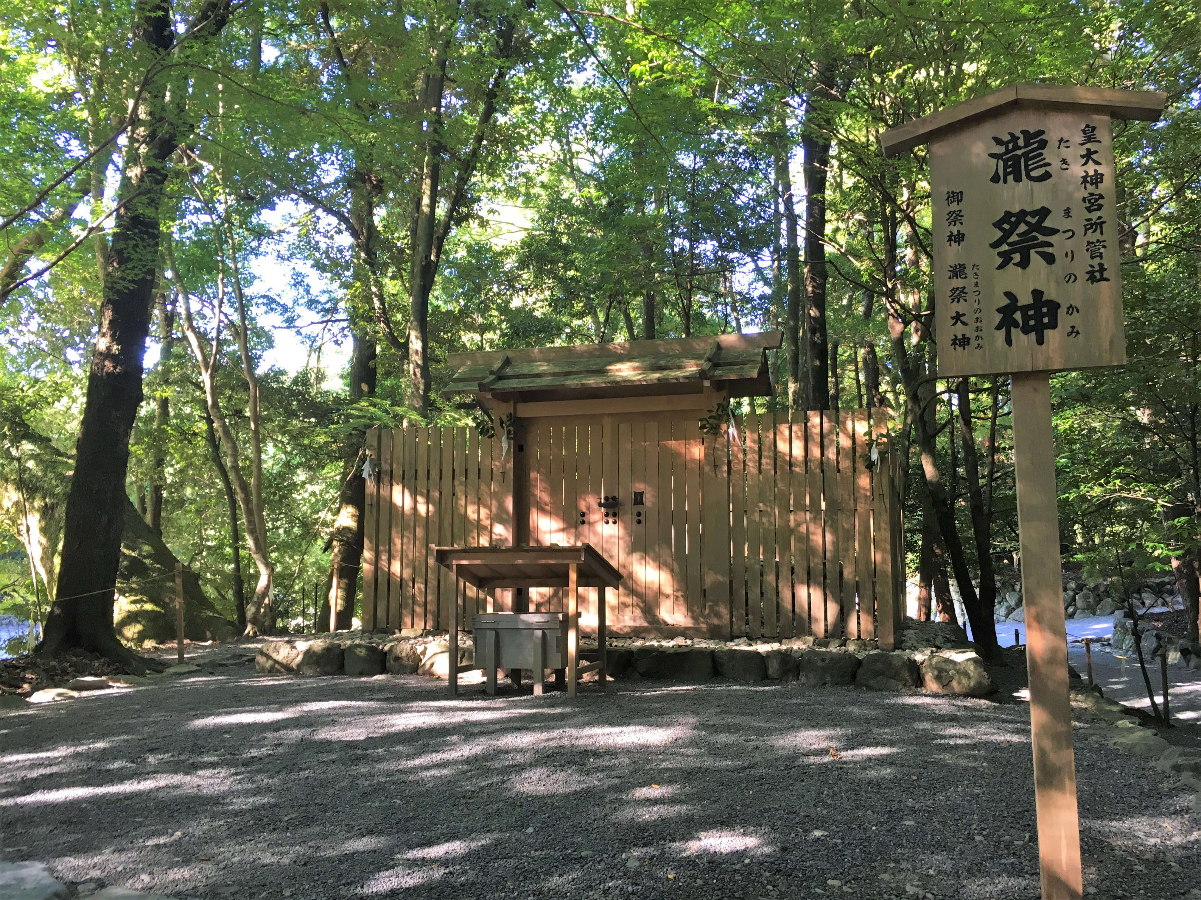 small wooden shinto shrine surrounded by a gravel yard and tall shady trees 