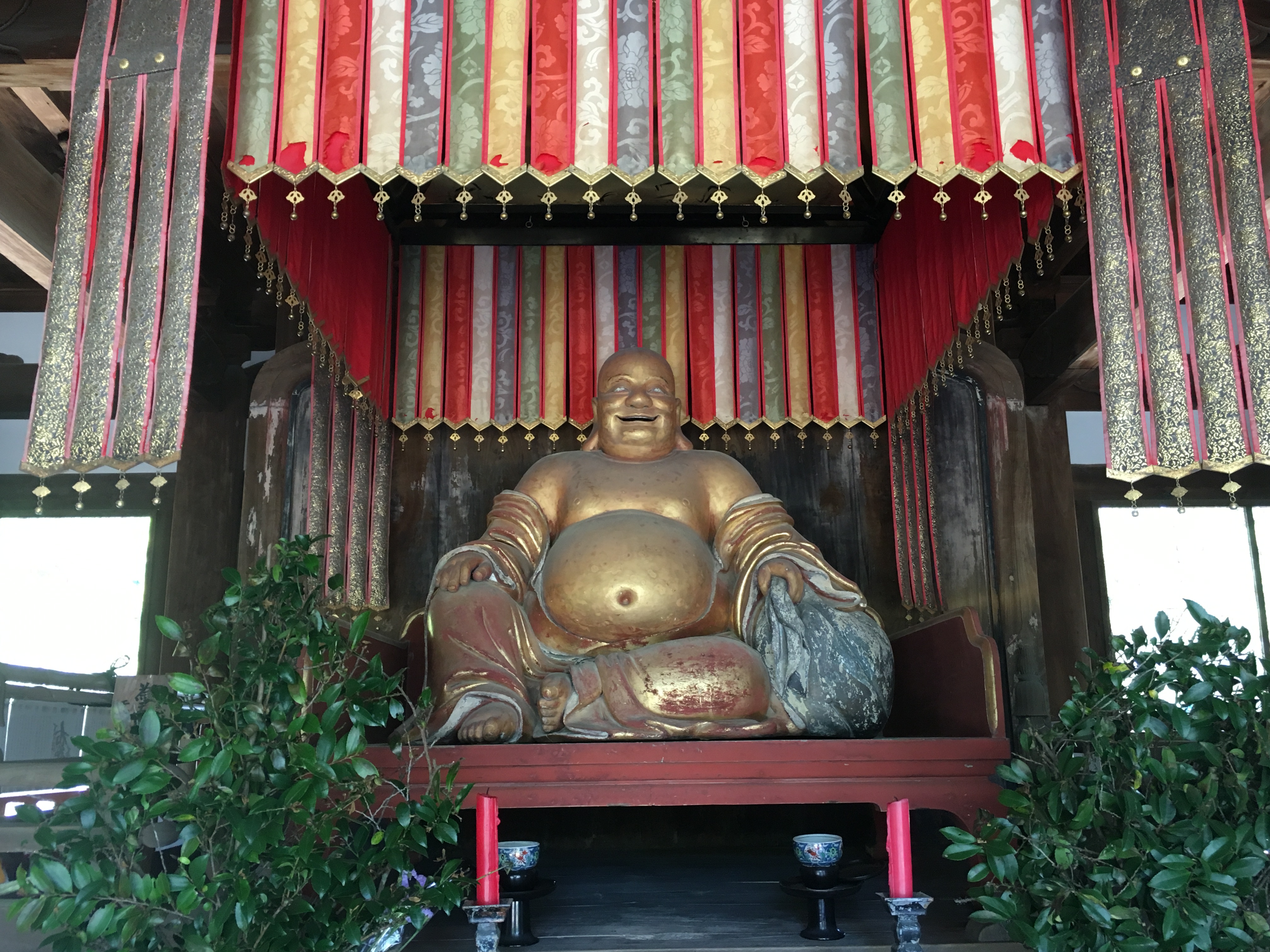 large golden statue of hotei in manpuku-ji temple under a rainbow of hanging fabric