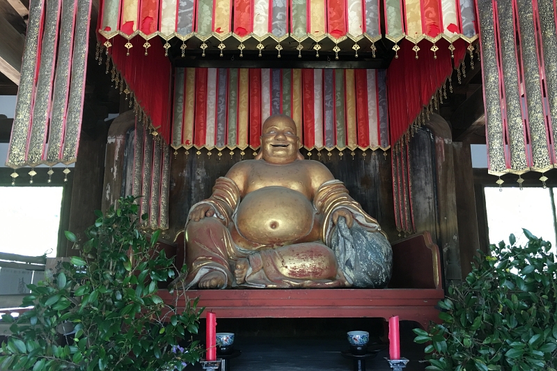 large golden statue of hotei in manpuku-ji temple under a rainbow of hanging fabric